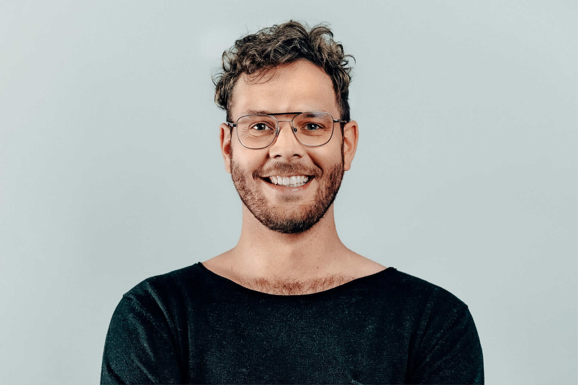 Ein freundlicher Mann mit Brille und dunklem Pullover, lächelnd, vor hellem Hintergrund.